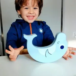 Uśmiechnięty chłopiec prezentuje drewnianą zabawkę w kształcie błękitnego wieloryba z rzędem kolorowych rybek na stole.
