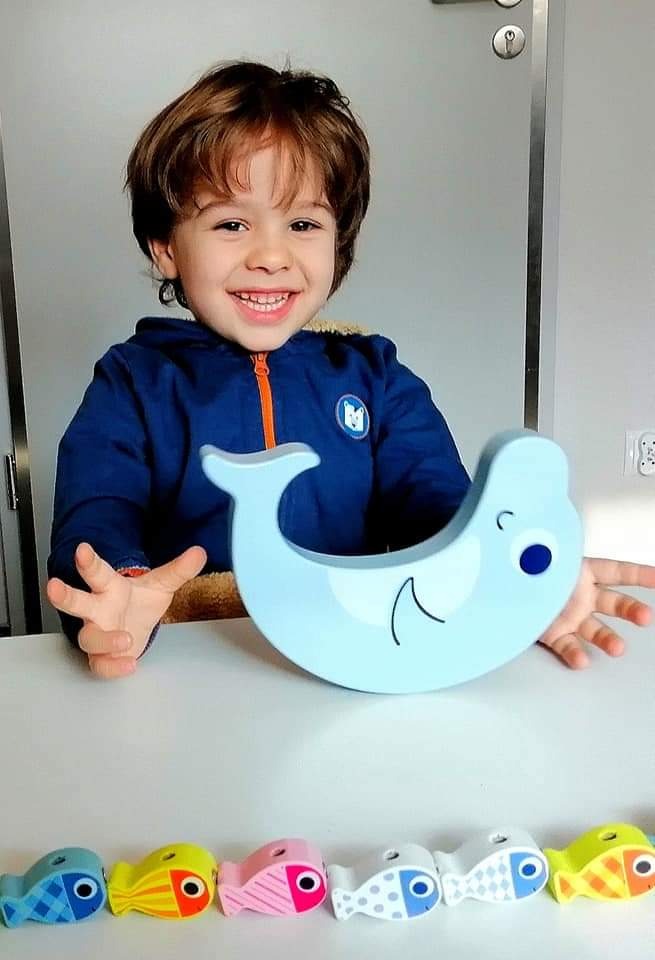 Uśmiechnięty chłopiec prezentuje drewnianą zabawkę w kształcie błękitnego wieloryba z rzędem kolorowych rybek na stole.