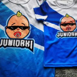 Dwie koszulki dziecięce z nadrukiem: jedna biało-błękitna, druga błękitna, obie z motywem niemowlaka w okularach przeciwsłonecznych i napisem 'JUNIORKI'.