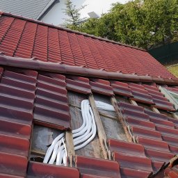 Gekon - Montaż instalacji wentylacyjnej z białymi rurami na dachu z czerwonej dachówki, widoczny fragment okna dachowego oraz samochód serwisowy z reklamą klimatyzacji w tle.