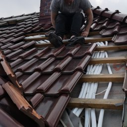 Gekon - Monter w szarej koszulce instaluje białe rury na dachu pokrytym brązową dachówką w pobliżu ceglanego komina i anteny.