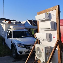 Gekon - Prezentacja klimatyzatorów różnych modeli na drewnianym stojaku z kółkami, obok samochód serwisowy z napisem 'Klimatyzacja Sprzedaż Montaż Serwis'.