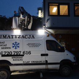 Gekon - Biały samochód serwisowy z podnośnikiem i napisem 'Klimatyzacja Sprzedaż Montaż Serwis' oświetlony reflektorami podczas pracy na zewnątrz budynku w nocy. Pracownik w podnośniku.