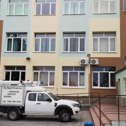 Gekon - Biały samochód serwisowy z napisem 'Klimatyzacja Sprzedaż Montaż Serwis' zaparkowany przed wielopiętrowym budynkiem z zainstalowanymi klimatyzatorami na zewnętrznej ścianie.