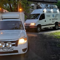 Gekon - Dwa samochody firmy klimatyzacyjnej, w tym jeden z podnośnikiem, zaparkowane na poboczu drogi w pobliżu domu, z włączonymi światłami ostrzegawczymi.