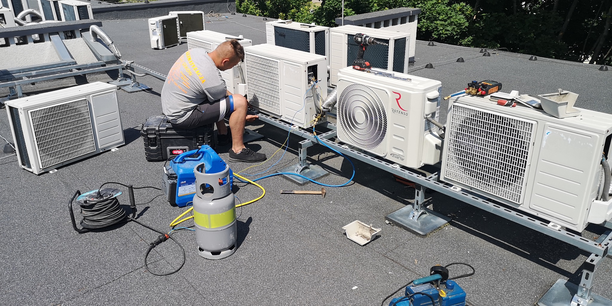 Instalator siedzi na czarnym kufrze narzędziowym i podłącza przewody do klimatyzatora Rotenso Inverter, na dachu budynku, z widocznymi butlami z gazem chłodniczym i narzędziami do serwisu...