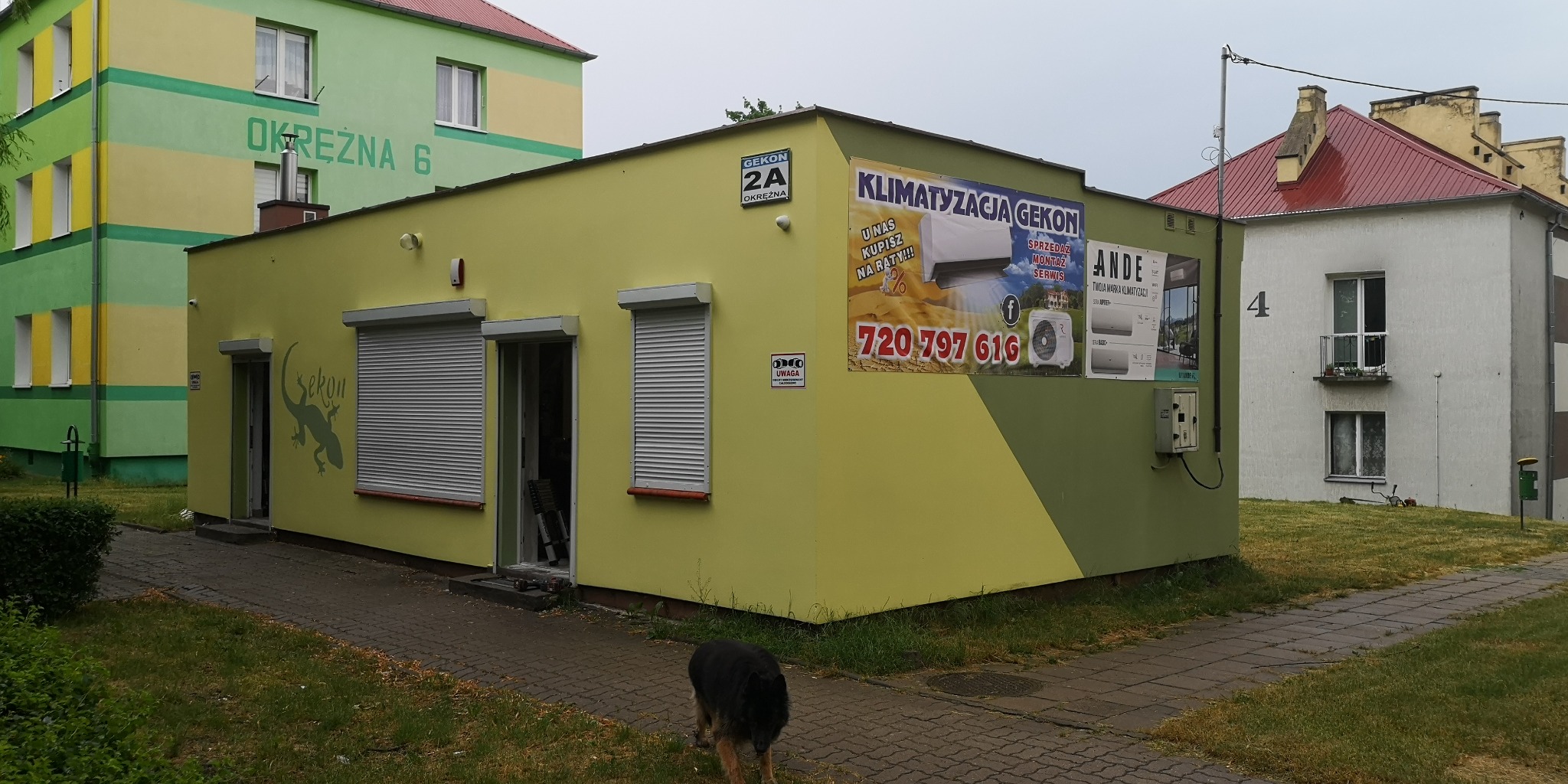 Zewnętrzny widok budynku firmy oferującej klimatyzację, z widocznymi reklamami na ścianie, żaluzjami w oknach i psem na chodniku przed budynkiem.