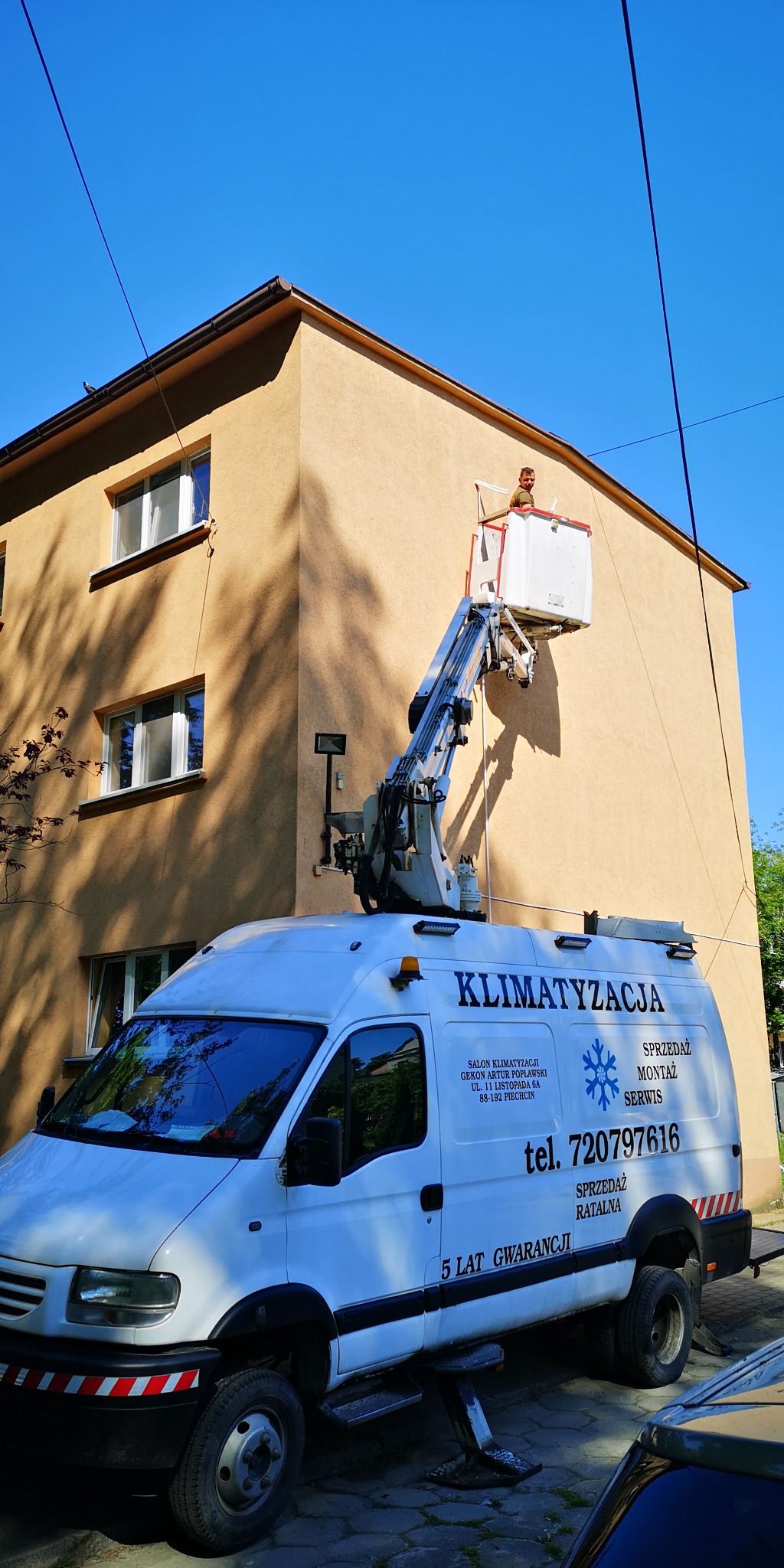 Pracownik na podnośniku koszowym montuje klimatyzację na ścianie budynku mieszkalnego, widoczny firmowy van z napisem 'Klimatyzacja' i numerem telefonu.