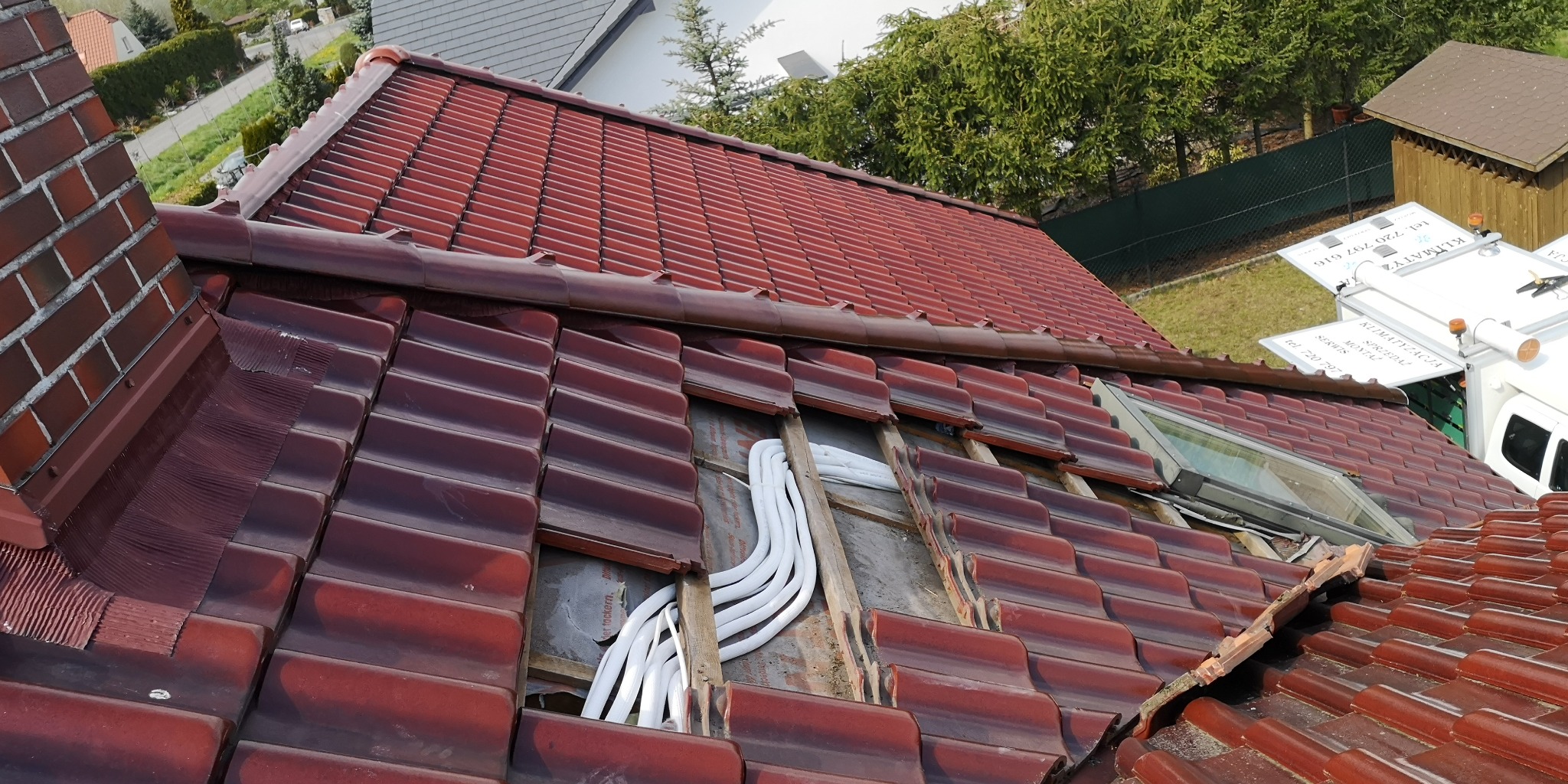 Montaż instalacji wentylacyjnej z białymi rurami na dachu z czerwonej dachówki, widoczny fragment okna dachowego oraz samochód serwisowy z reklamą klimatyzacji w tle.