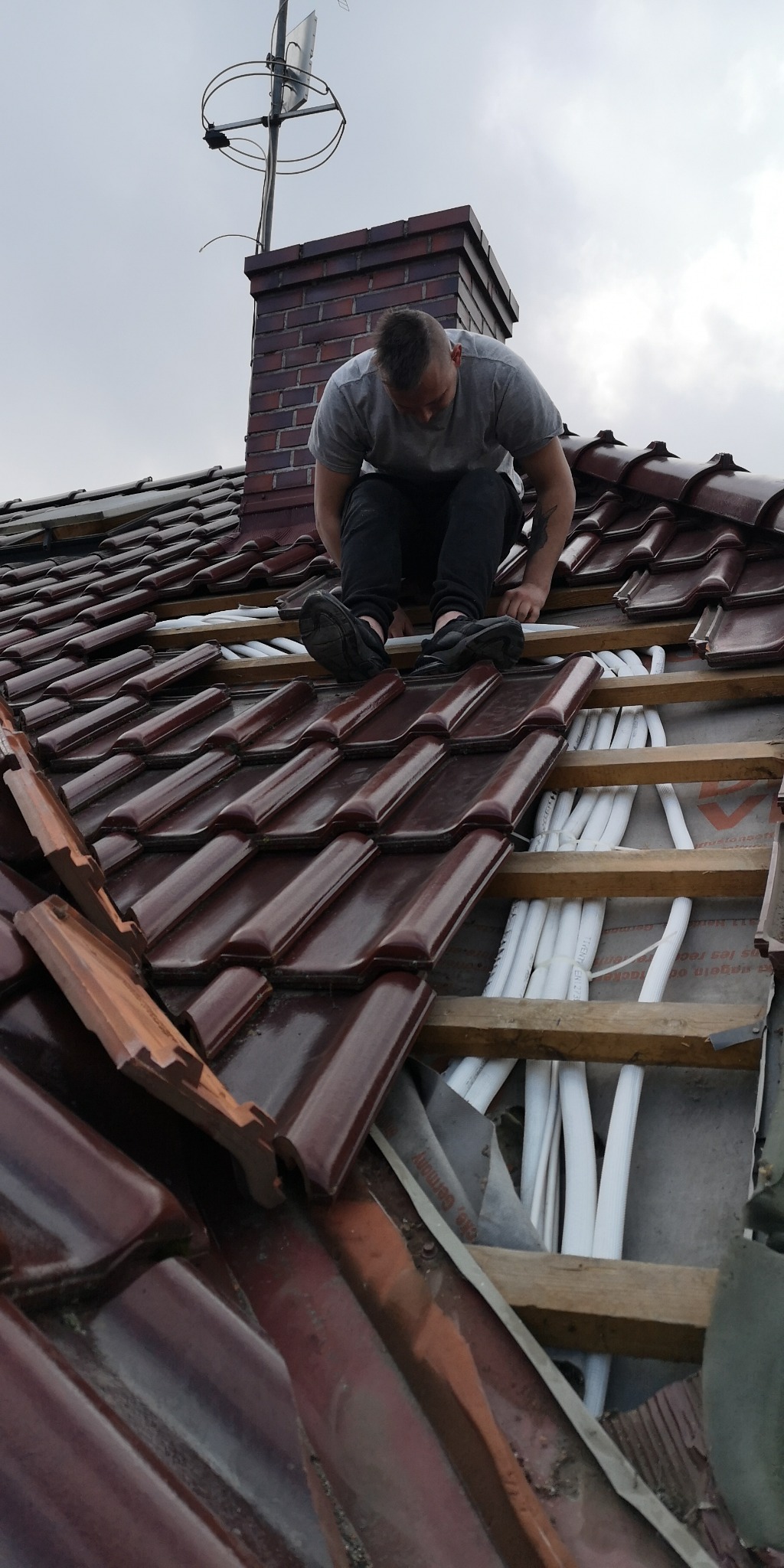 Monter w szarej koszulce instaluje białe rury na dachu pokrytym brązową dachówką w pobliżu ceglanego komina i anteny.