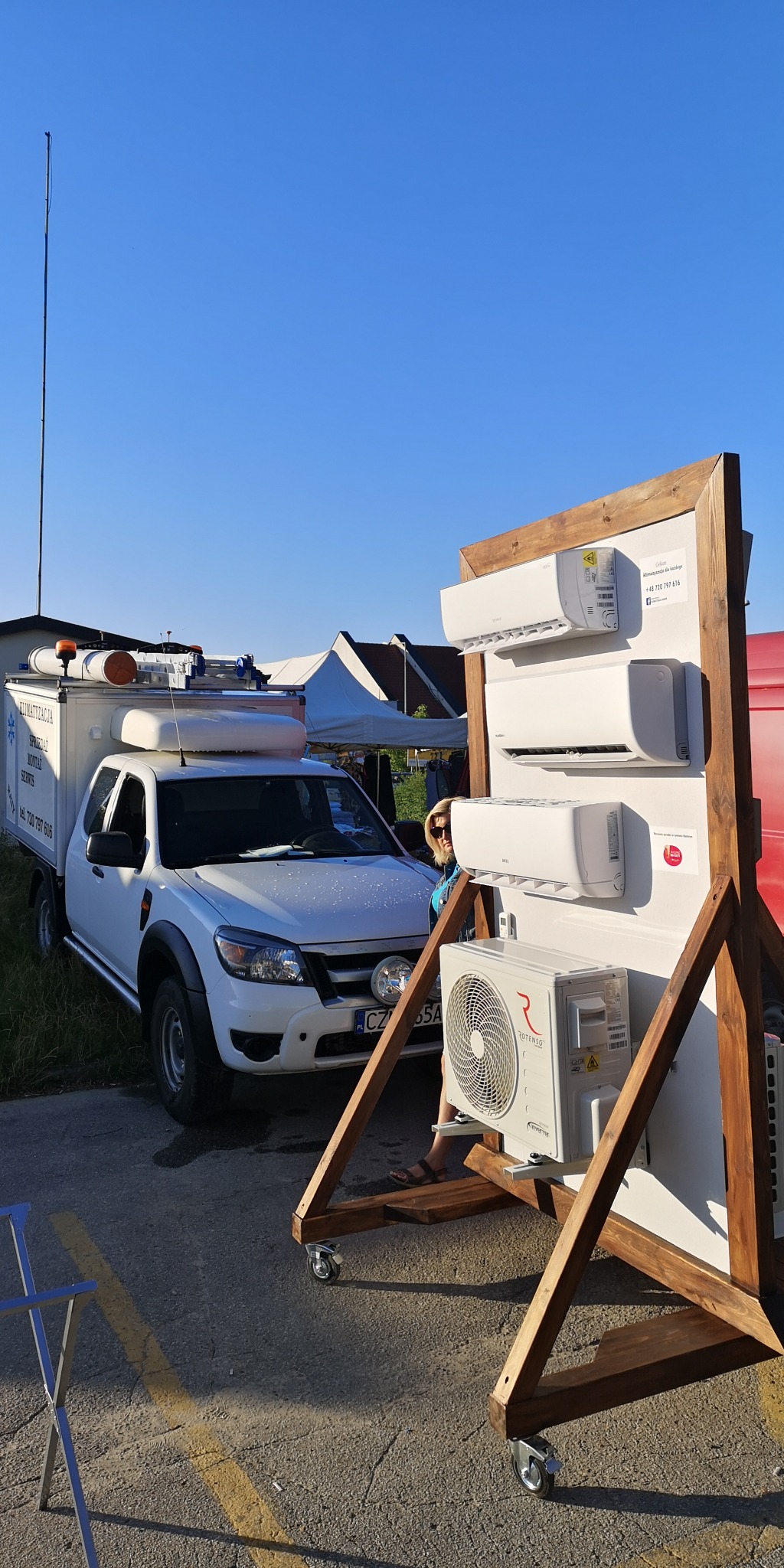 Prezentacja klimatyzatorów różnych modeli na drewnianym stojaku z kółkami, obok samochód serwisowy z napisem 'Klimatyzacja Sprzedaż Montaż Serwis'.