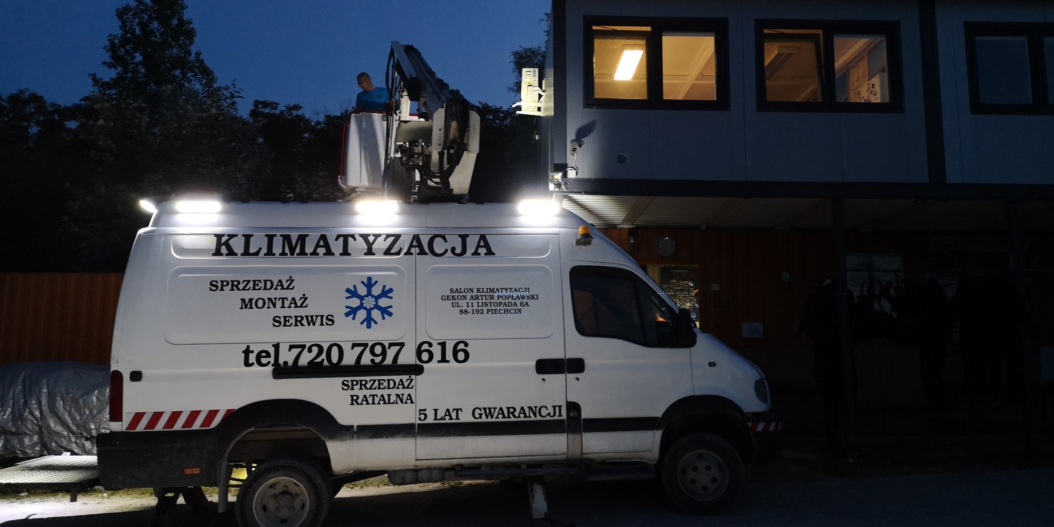Biały samochód serwisowy z podnośnikiem i napisem 'Klimatyzacja Sprzedaż Montaż Serwis' oświetlony reflektorami podczas pracy na zewnątrz budynku w nocy. Pracownik w podnośniku.