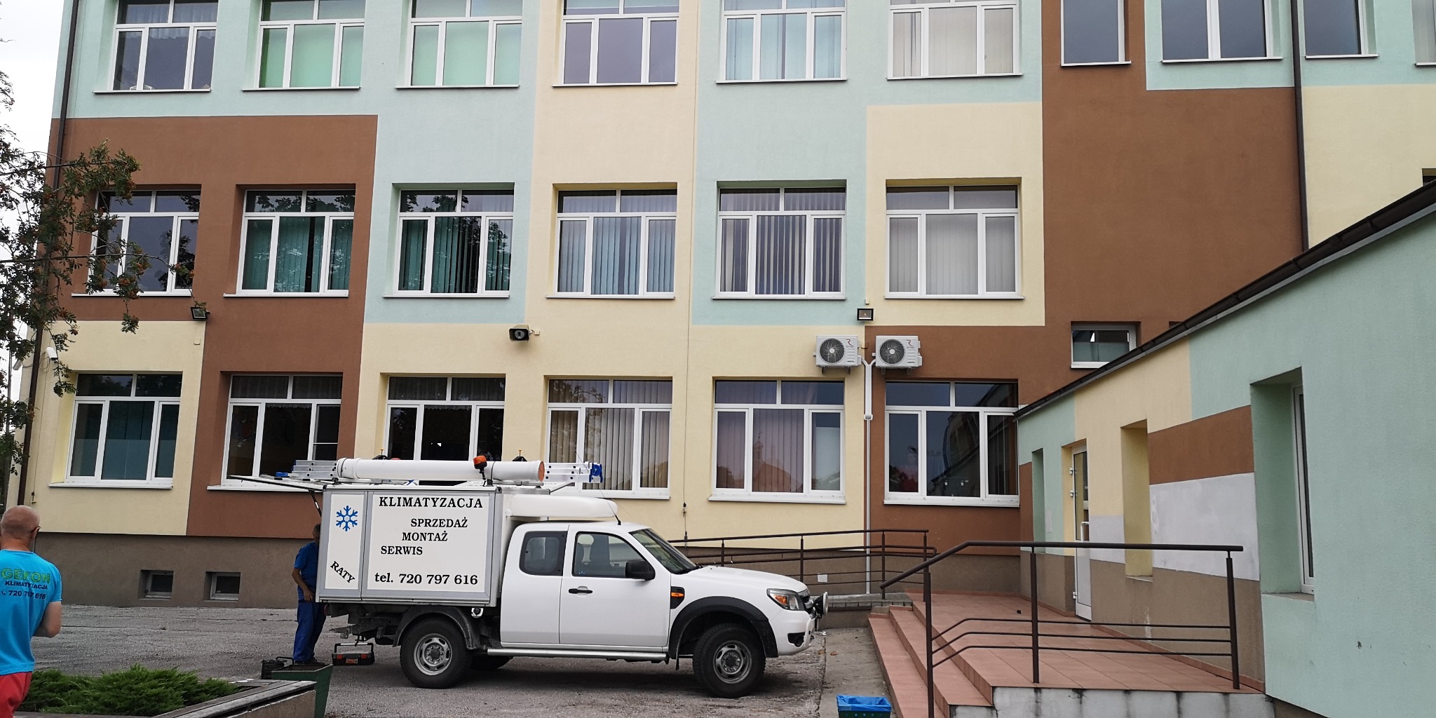 Biały samochód serwisowy z napisem 'Klimatyzacja Sprzedaż Montaż Serwis' zaparkowany przed wielopiętrowym budynkiem z zainstalowanymi klimatyzatorami na zewnętrznej ścianie.