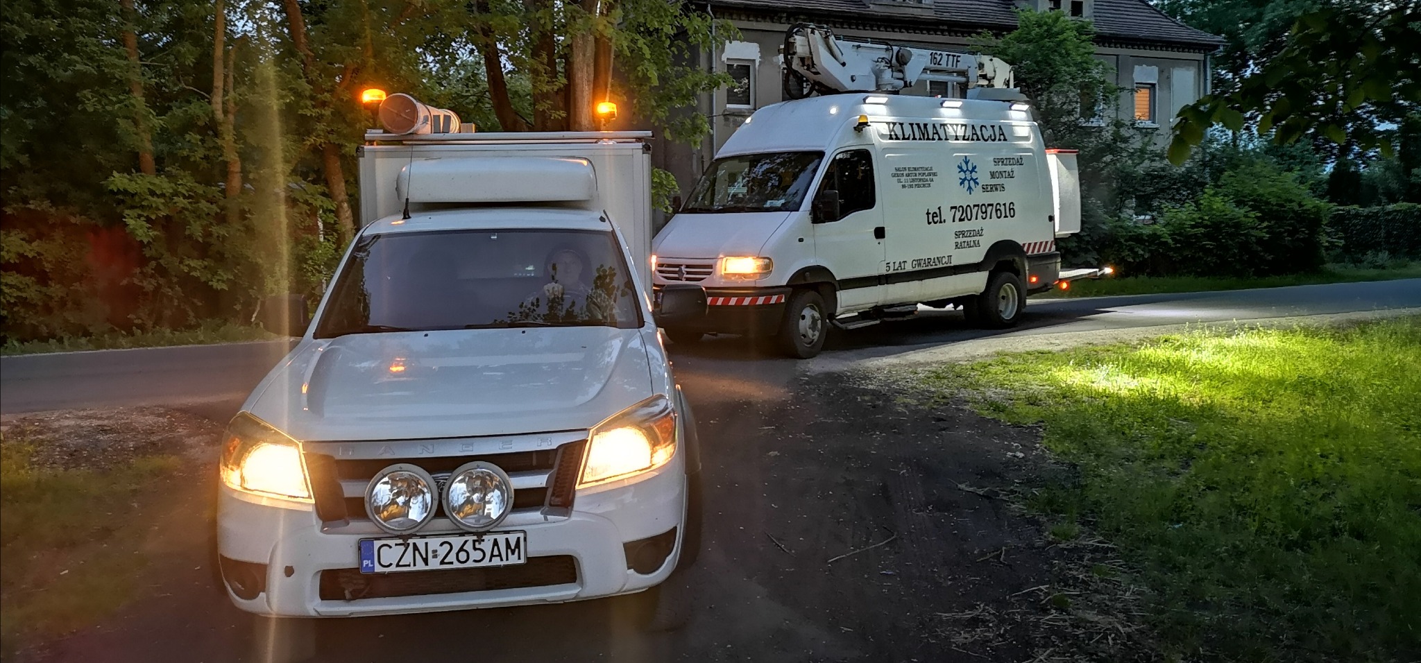 Dwa samochody firmy klimatyzacyjnej, w tym jeden z podnośnikiem, zaparkowane na poboczu drogi w pobliżu domu, z włączonymi światłami ostrzegawczymi.