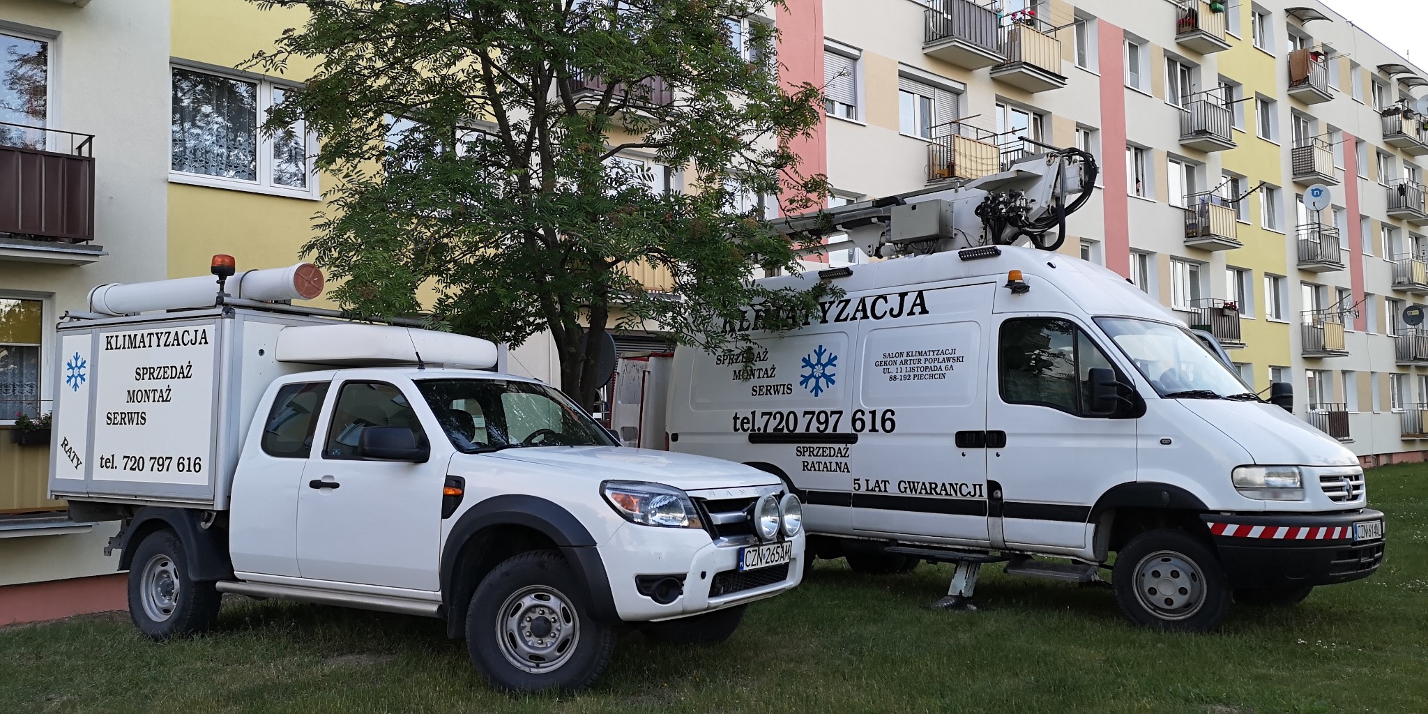 Dwa białe samochody serwisowe z napisami 'Klimatyzacja Sprzedaż Montaż Serwis' zaparkowane na trawniku przed blokiem mieszkalnym. Jeden samochód to pickup z zabudową, drugi to furgonetka...