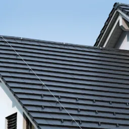 Widok z dołu na dwa fragmenty dachu pokryte ciemnoszarą dachówką ceramiczną, z widocznym systemem odgromowym i fragmentem białej elewacji budynku.