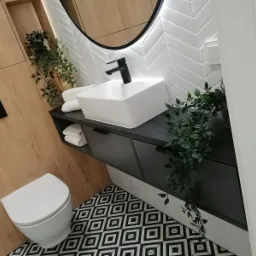 Nowoczesna łazienka z umywalką nablatową, czarną armaturą, szafką pod umywalką z szufladami, lustrem z podświetleniem LED, białą mozaiką w jodełkę na ścianie, drewnianą okładziną ścienną i podłogą...