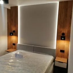 Nowoczesna sypialnia z drewnianymi panelami oświetlonymi LED po bokach łóżka, minimalistyczne lampki nocne i szafki, łóżko zabezpieczone folią ochronną, pościel w pudełku na łóżku.