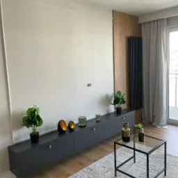 Nowoczesny salon z szarą, podwieszaną szafką RTV z dekoracjami, drewnianą ścianą i czarnym grzejnikiem.