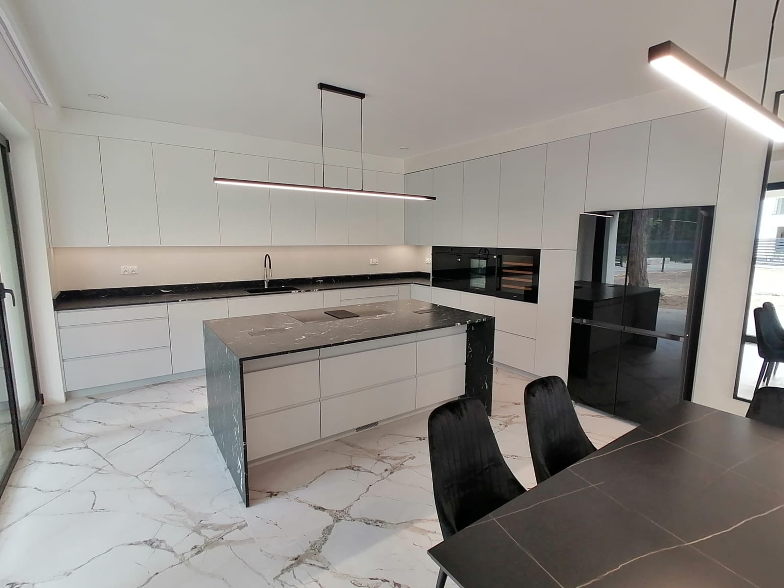 Nowoczesna kuchnia z wyspą, białe szafki, czarny blat z marmurowym wzorem, oświetlenie LED, podłoga z białego marmuru.