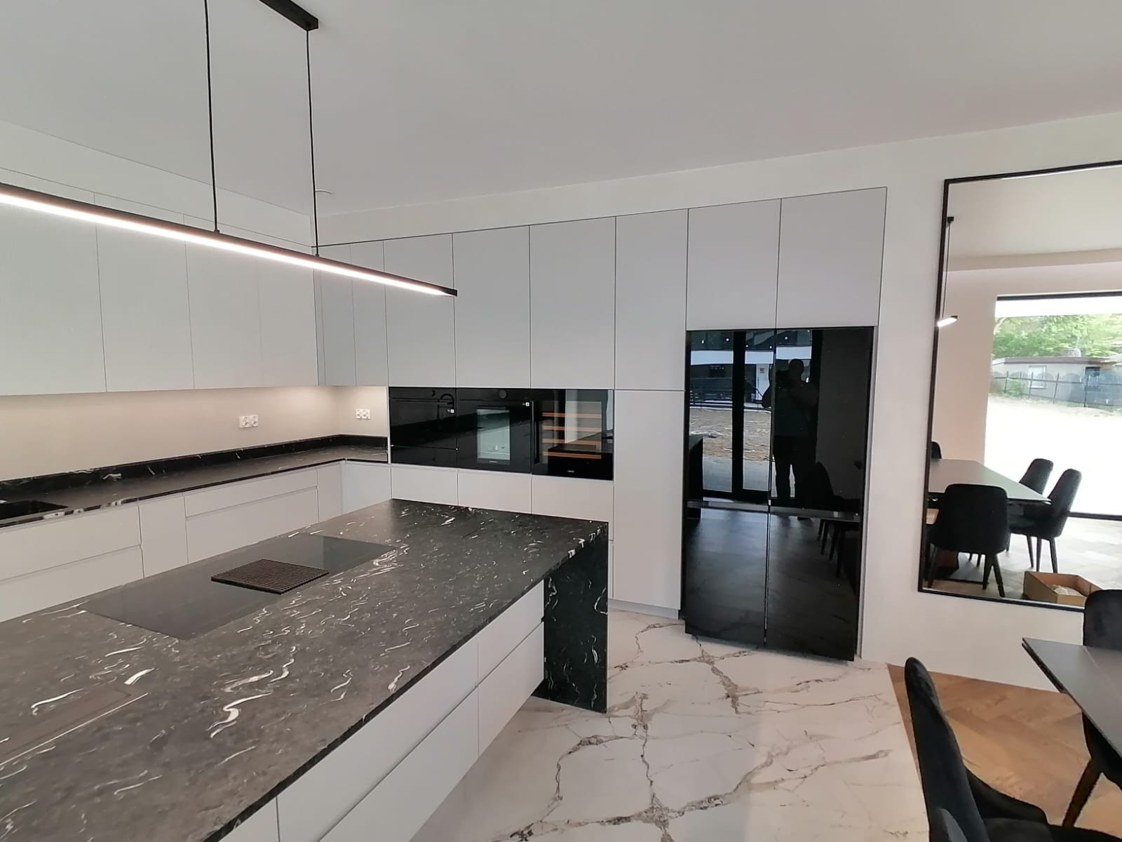 Nowoczesna kuchnia w Woli Jedlińskiej z białymi szafkami, czarnym blatem z marmurowym wzorem, podłużną lampą sufitową i podłogą z białego marmuru z ciemnymi żyłami, z widoczną zabudową sprzętów AGD.