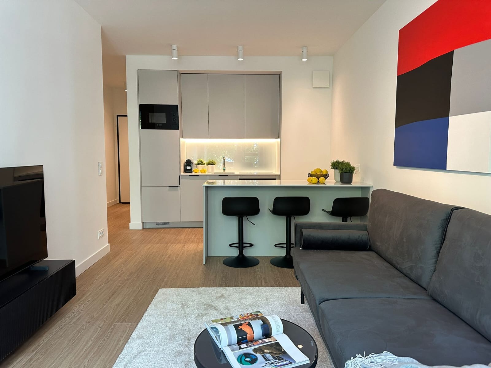 Nowoczesna kuchnia w odcieniach szarości z wyspą i czarnymi hokerami, widok z salonu z szarą kanapą i abstrakcyjnym obrazem na ścianie.