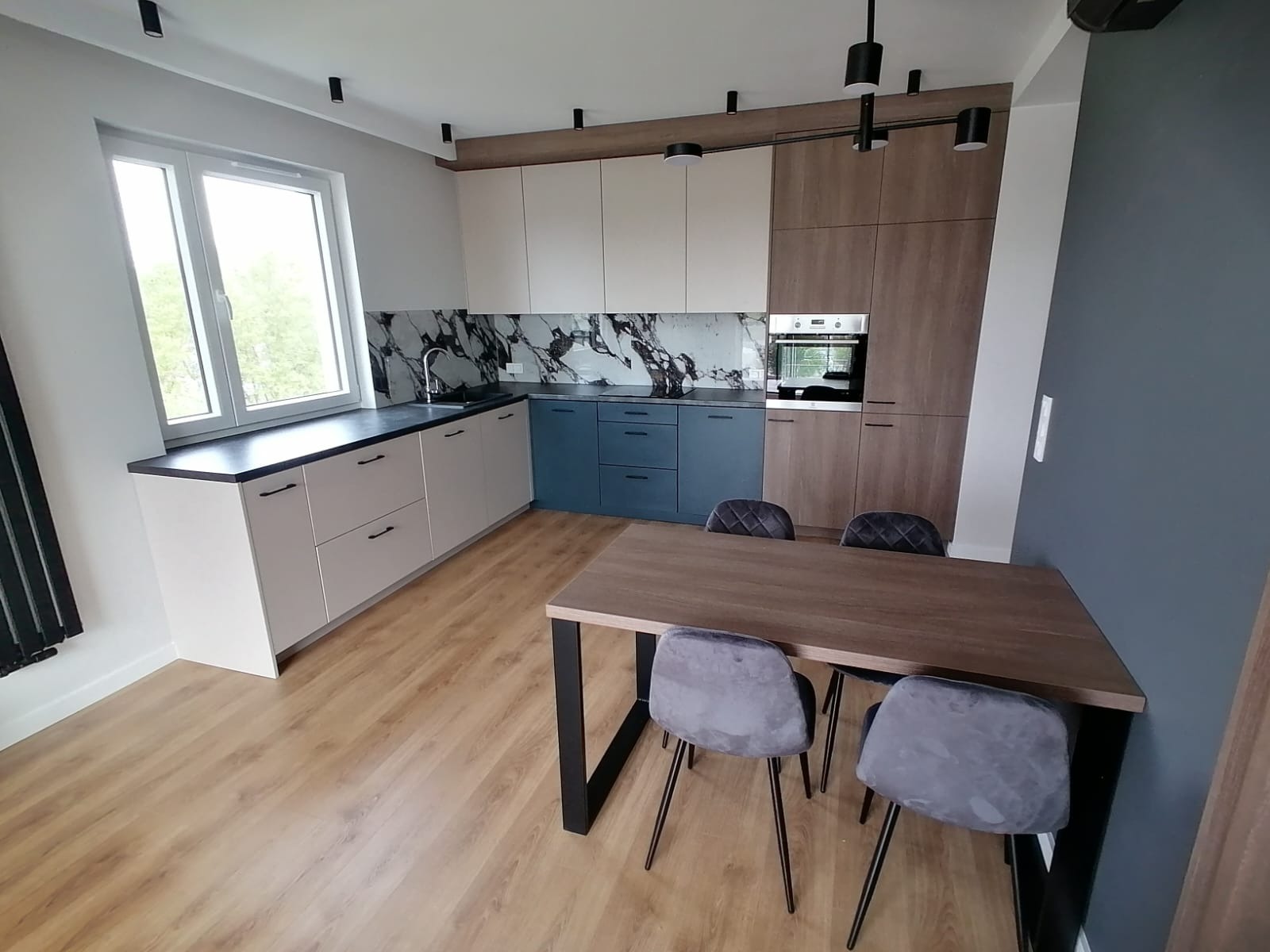 Nowoczesna kuchnia z zabudową w odcieniach beżu, błękitu i drewna, marmurowy backsplash, czarny blat i oświetlenie punktowe, stół z szarymi krzesłami