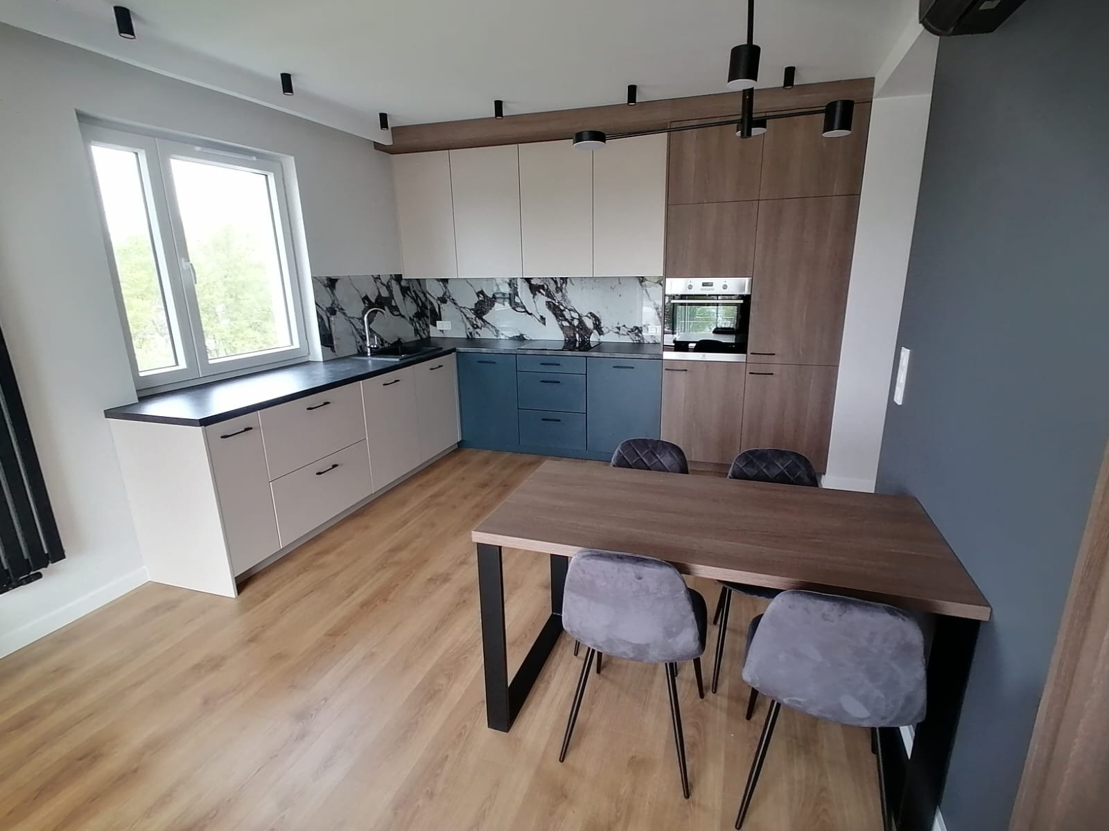 Nowoczesna kuchnia z zabudową w odcieniach beżu, błękitu i drewna, z blatem imitującym marmur i drewnianym stołem jadalnianym z szarymi krzesłami.