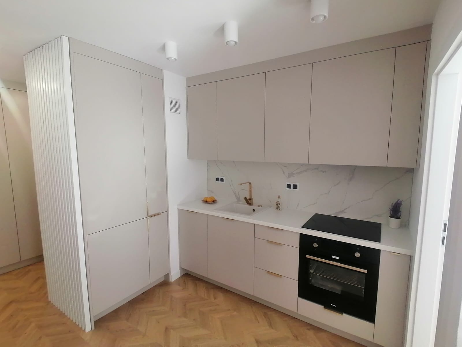 Jasna kuchnia z zabudową pod sufit, fronty w kolorze beżowym, złota armatura, marmurowy backsplash i parkiet w jodełkę.