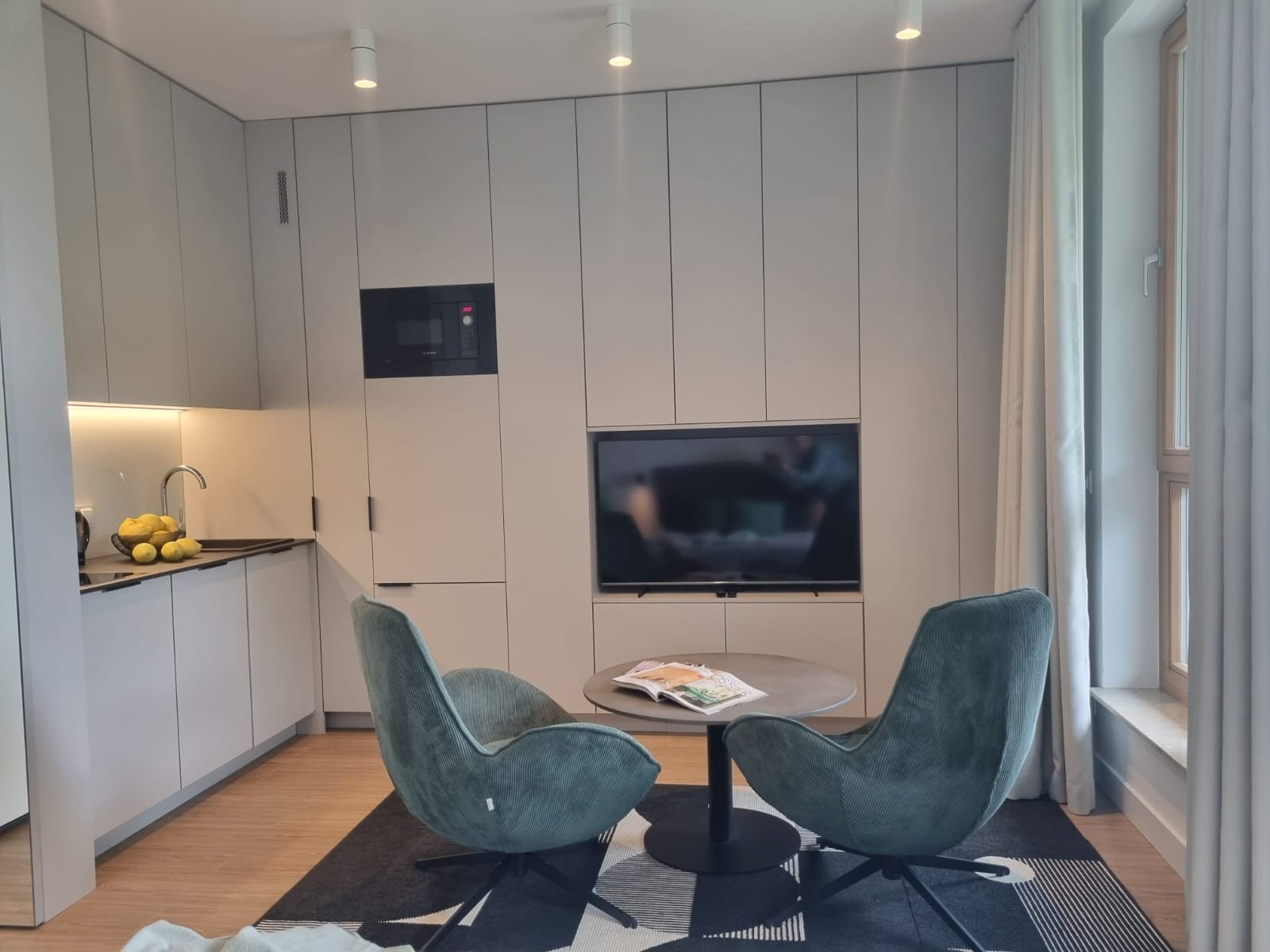 Nowoczesna, jasna kuchnia z zabudową meblową sięgającą sufitu, zintegrowaną z szafkami RTV, z okrągłym stołem i dwoma zielonymi fotelami na tle wzorzystego dywanu.