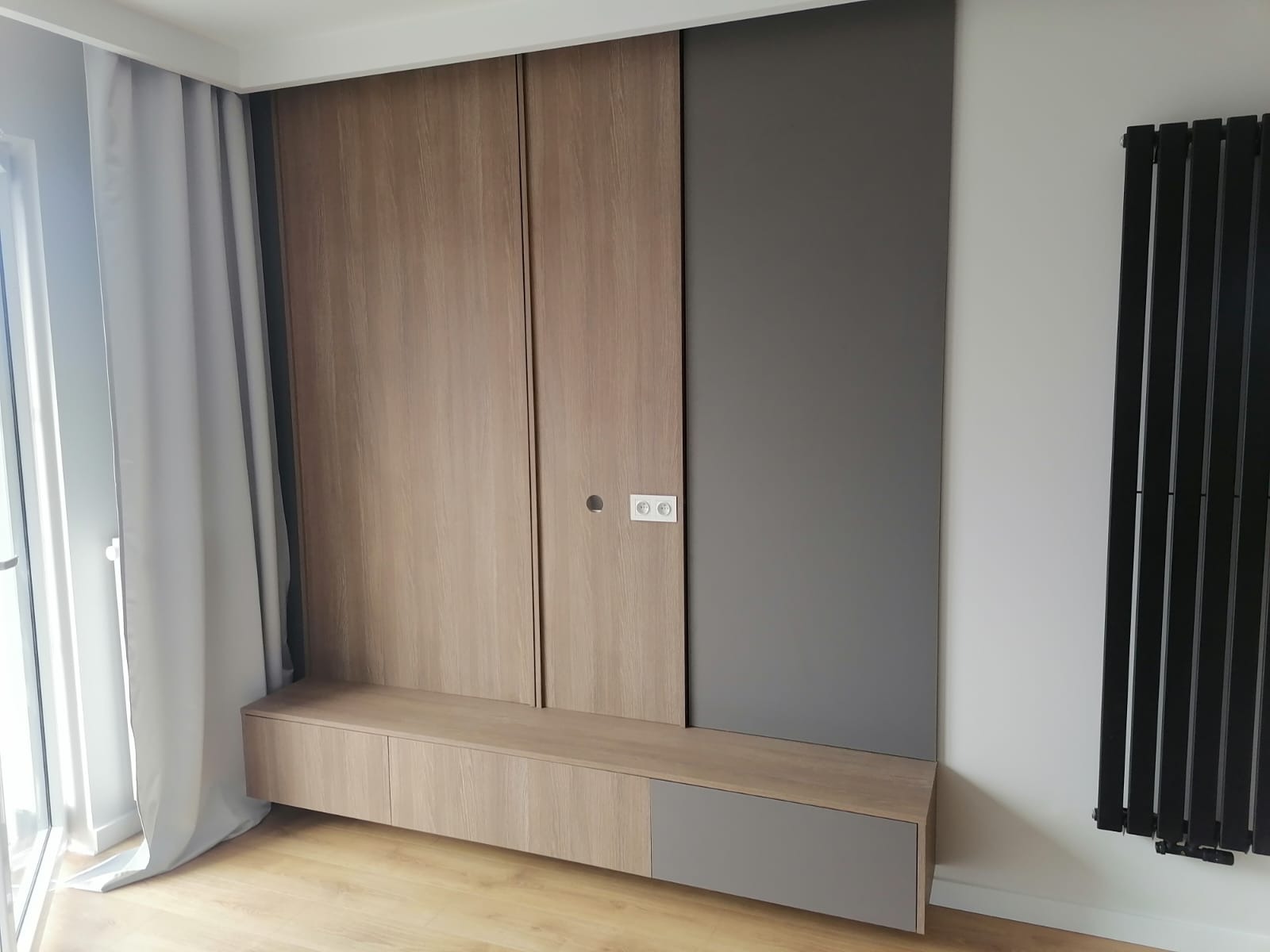Szafa wnękowa z przesuwnymi drzwiami w odcieniach drewna i szarości, zintegrowana z niską komodą, obok grzejnik panelowy w kolorze czarnym.