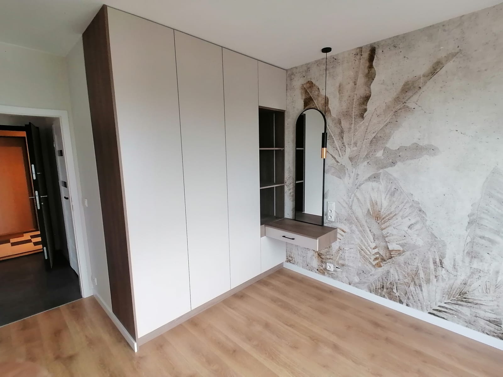 Biała szafa wnękowa z drewnianymi akcentami, toaletka z lustrem w czarnej ramie, ściana z motywem liści, jasna podłoga z paneli.