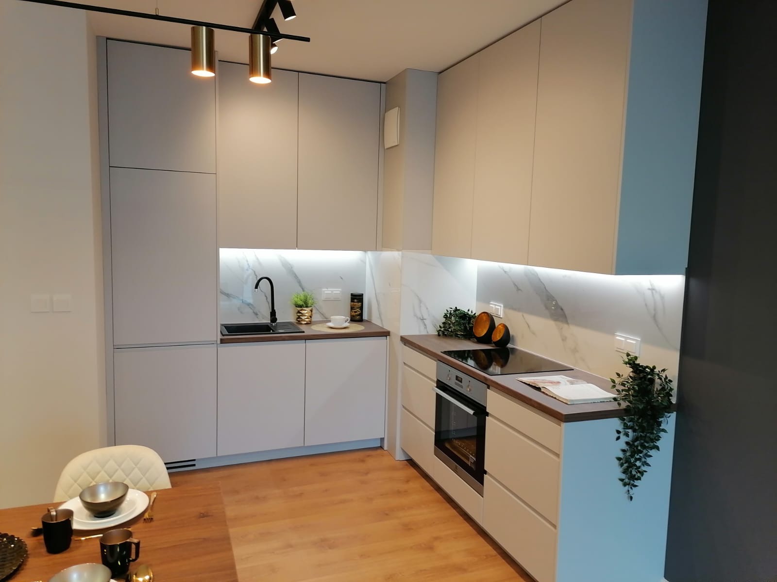 Jasnoszara kuchnia z marmurowym backsplashiem i czarną armaturą, oświetlenie punktowe na suficie, stół z zastawą w pierwszym planie.