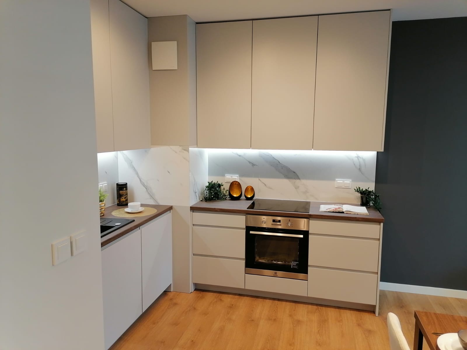 Jasna kuchnia z białymi frontami, marmurowym backsplashiem podświetlonym LED, drewnianym blatem, piekarnikiem i dekoracyjnymi wazonami.