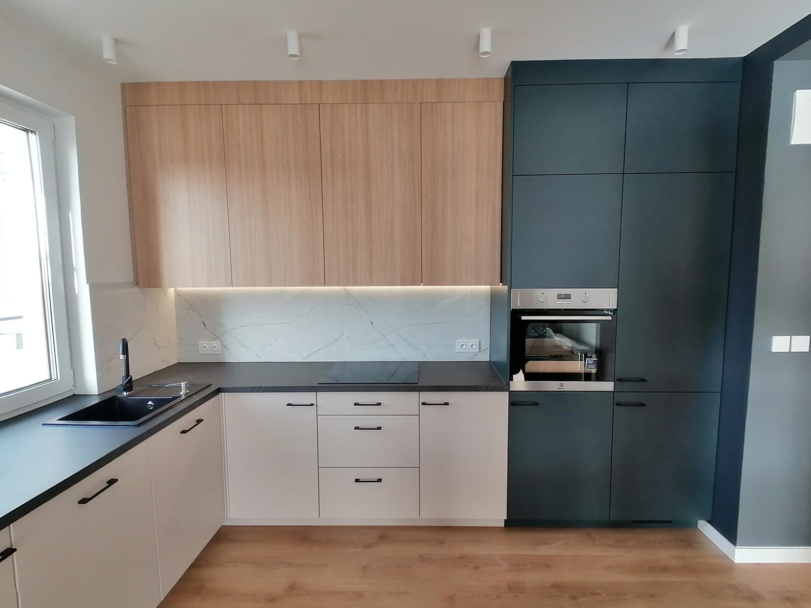 Nowoczesna kuchnia z drewnianymi górnymi szafkami, białymi dolnymi szafkami i ciemnogranatową wysoką zabudową z piekarnikiem, podświetlony blat z marmurowym panelem.