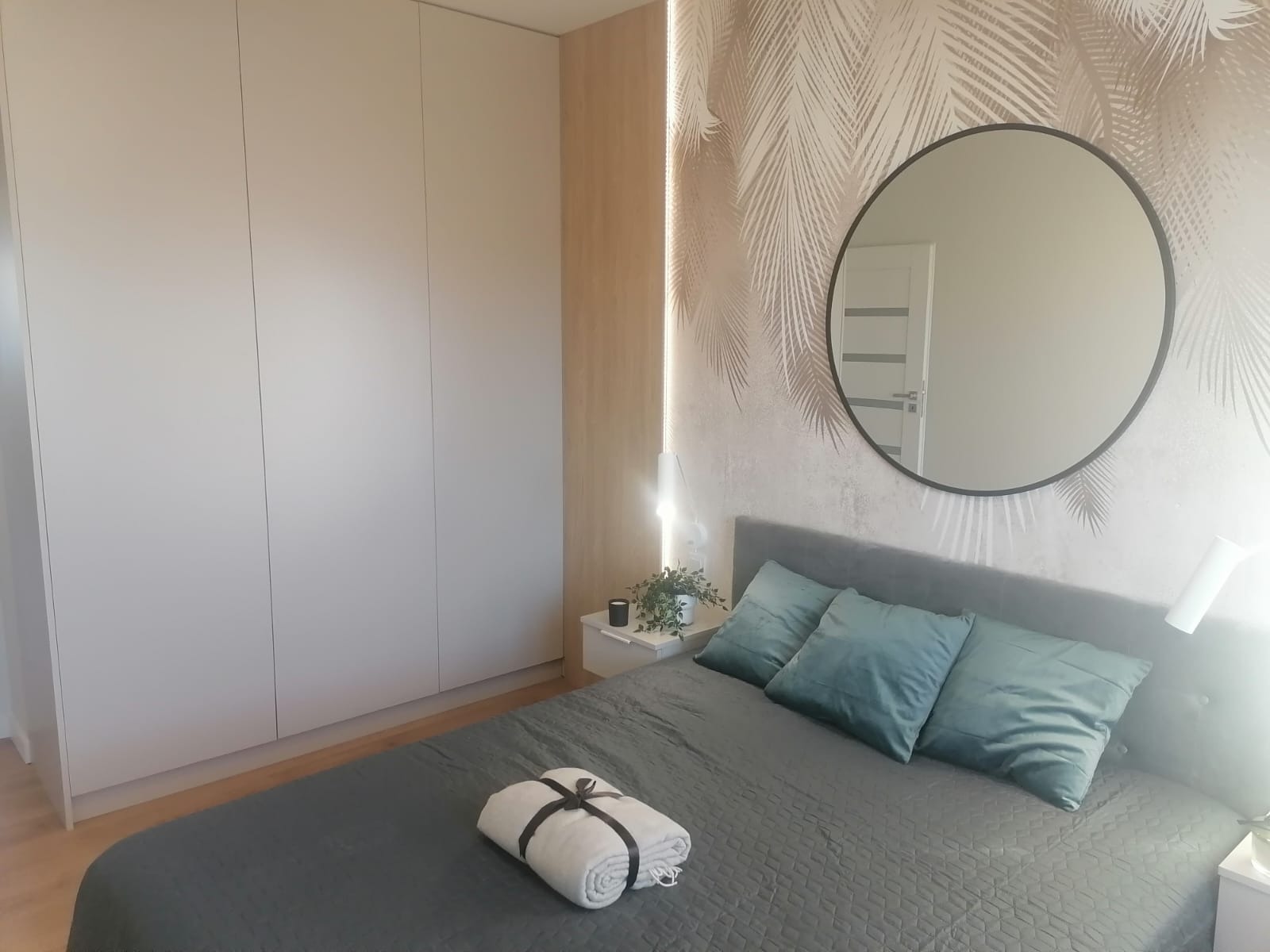 Sypialnia z dużymi, białymi szafami na wymiar, okrągłym lustrem na ścianie w palmy i szarym łóżkiem z turkusowymi poduszkami.