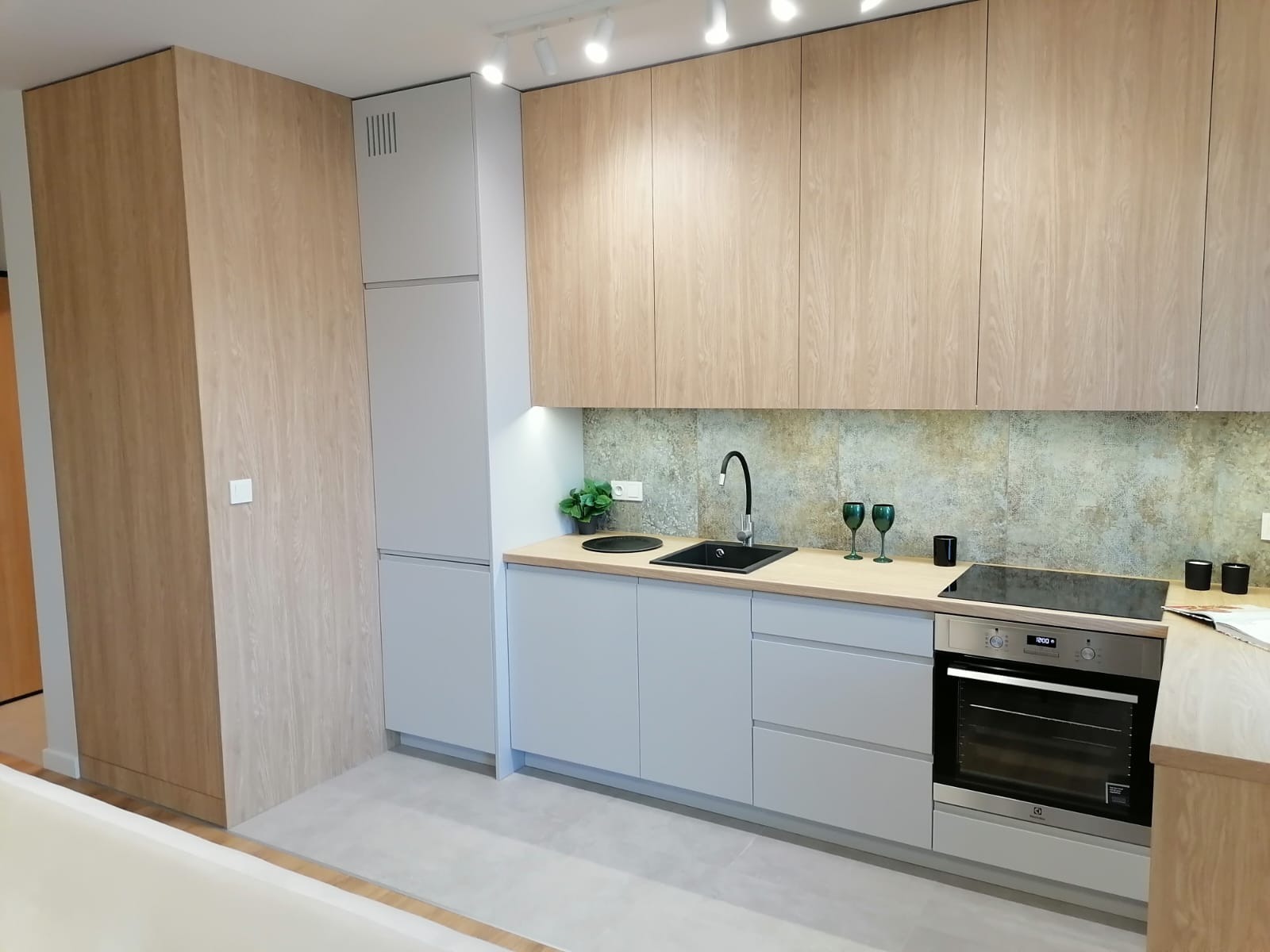 Jasna kuchnia z drewnianymi szafkami górnymi i szarymi dolnymi, czarny zlew i bateria, zielone szklanki na blacie, oświetlenie punktowe.