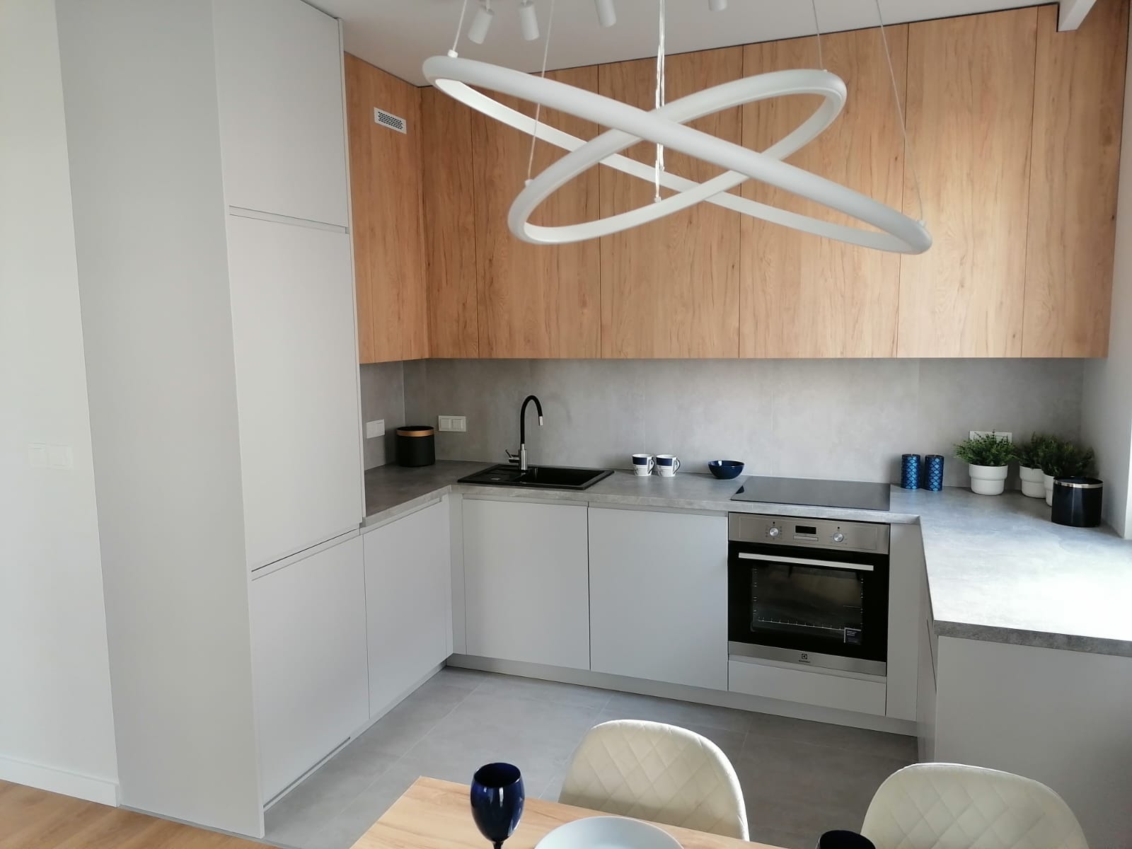 Nowoczesna kuchnia z białymi frontami i drewnianymi szafkami górnymi, oświetlona designerską lampą pierścieniową, z widocznym fragmentem stołu jadalnego z nakryciem.