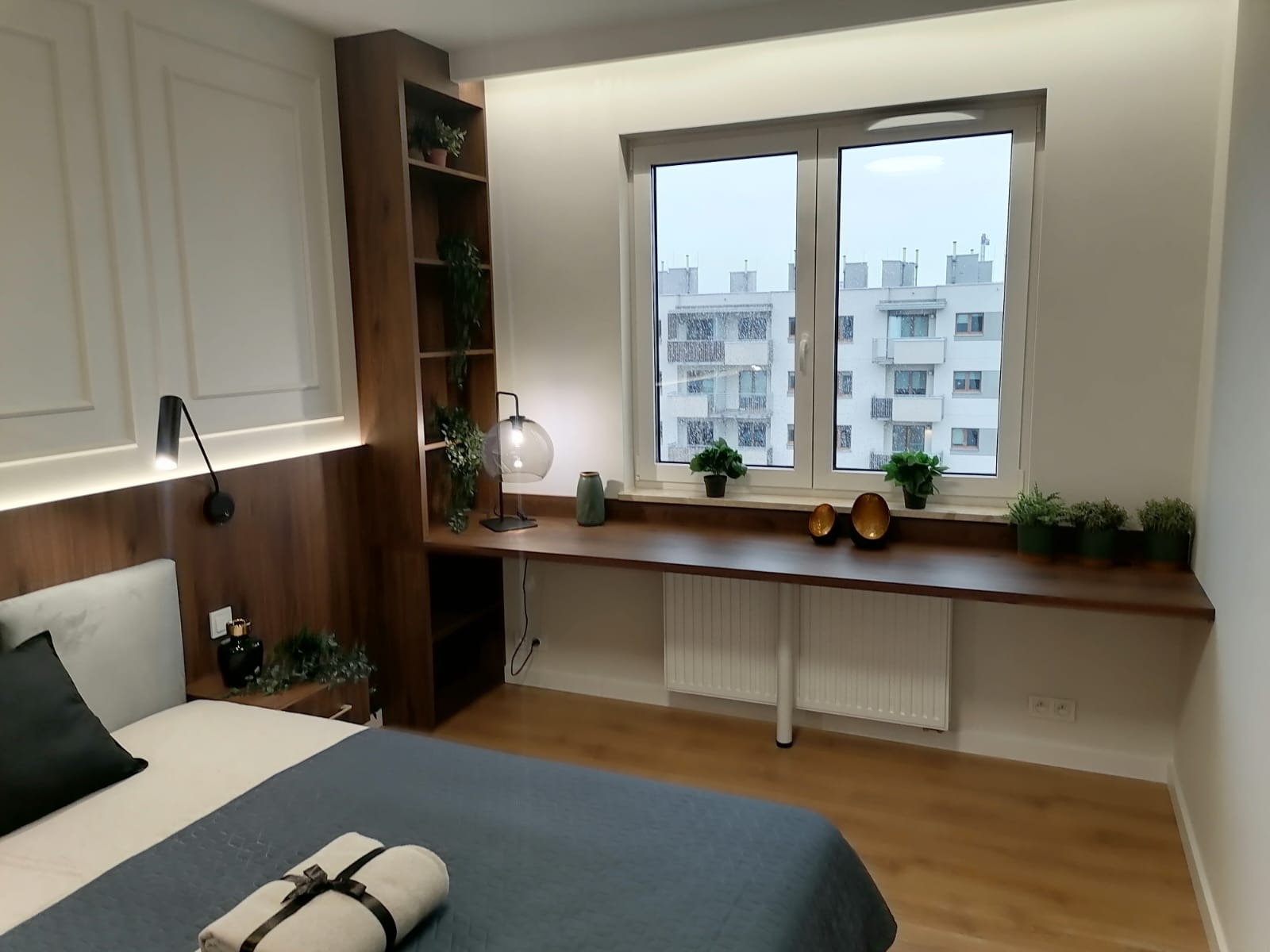 Nowoczesna sypialnia z drewnianą zabudową, biurkiem pod oknem i półkami z roślinami, widok na budynek mieszkalny za oknem.