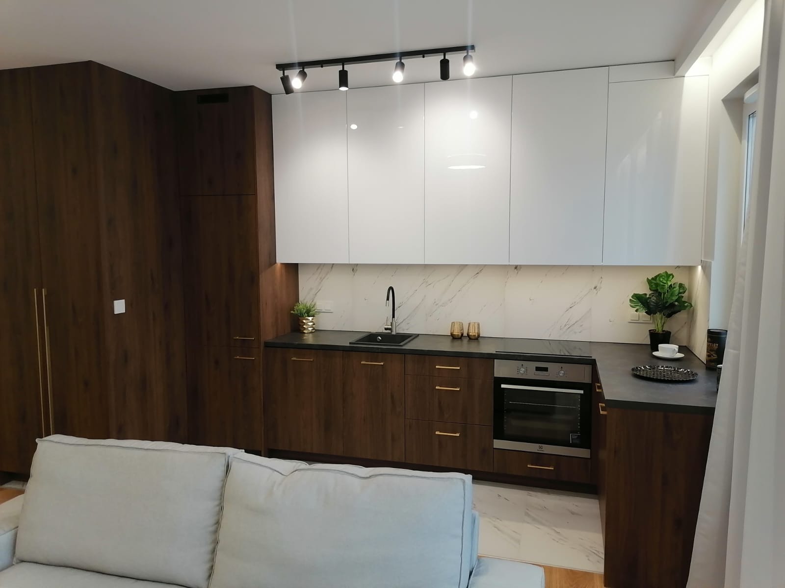 Nowoczesna kuchnia z białymi szafkami górnymi i dolnymi w odcieniach brązu z drewnianą fakturą, czarny blat, marmurowa ściana nad blatem, oświetlenie punktowe i fragment szarej sofy.
