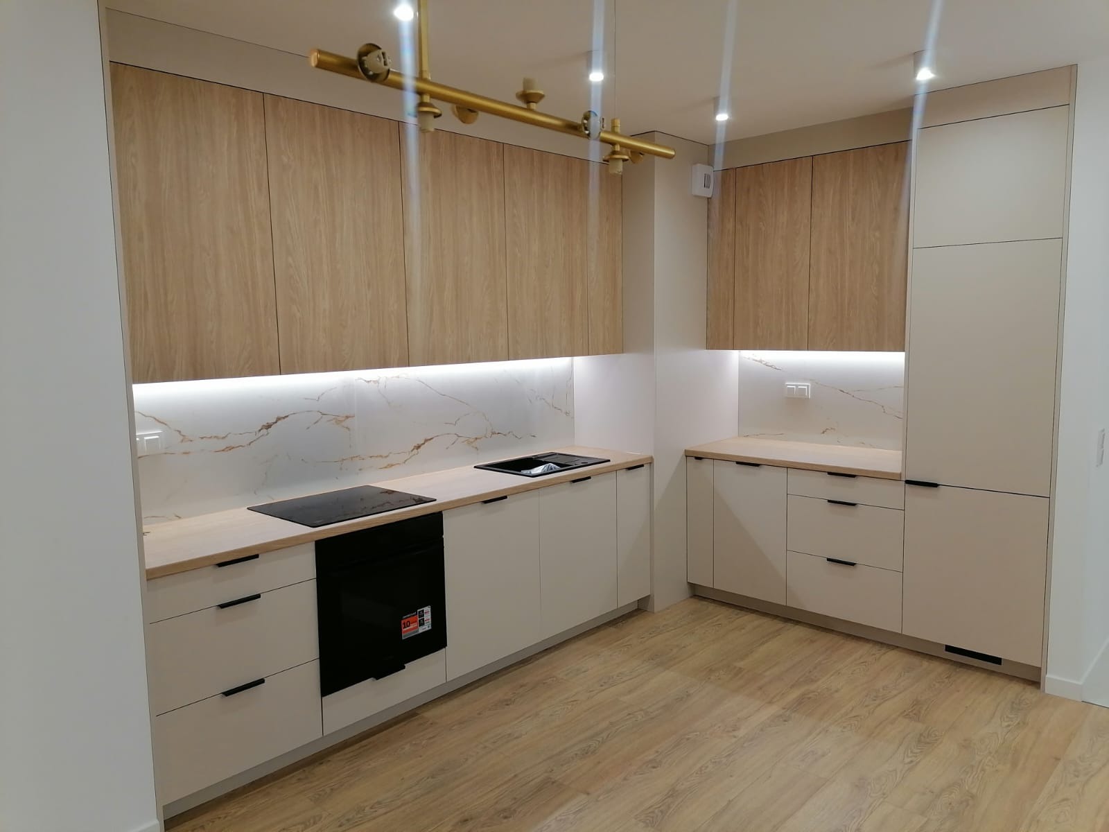 Nowoczesna kuchnia z białymi szafkami dolnymi, drewnopodobnymi szafkami górnymi, blatem w kolorze jasnego drewna i marmurowym backsplashiem podświetlonym listwą LED, złoty żyrandol.