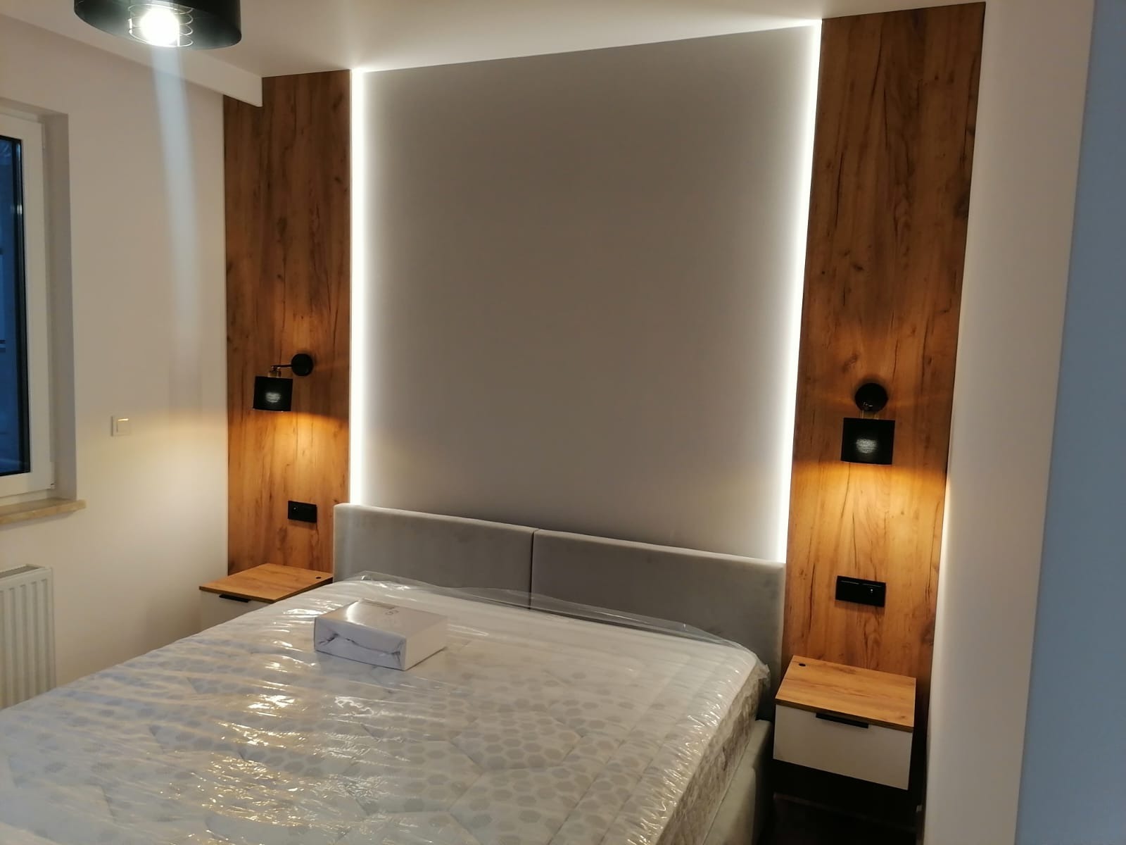 Nowoczesna sypialnia z drewnianymi panelami oświetlonymi LED po bokach łóżka, minimalistyczne lampki nocne i szafki, łóżko zabezpieczone folią ochronną, pościel w pudełku na łóżku.