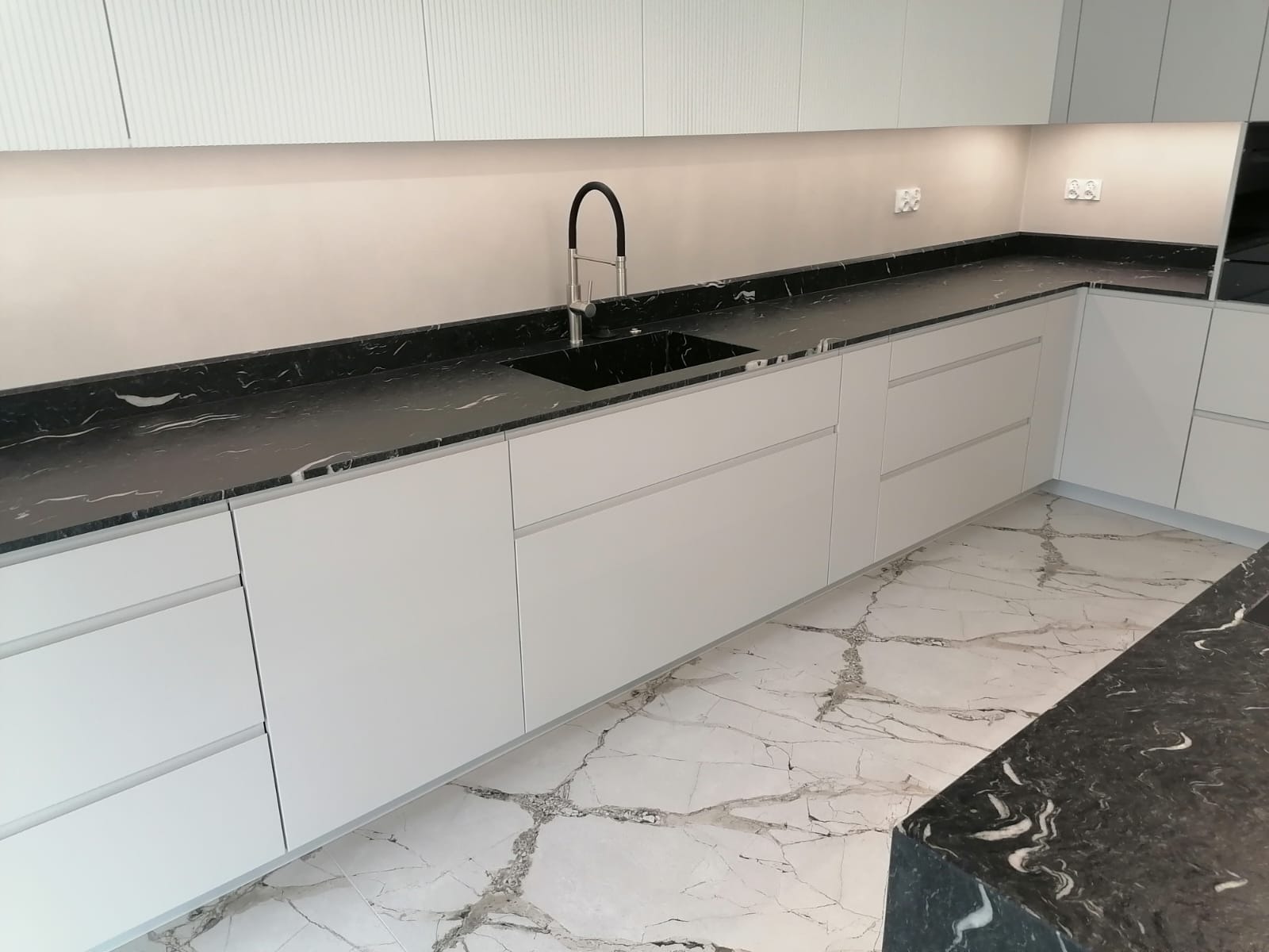 Nowoczesna, minimalistyczna kuchnia z białymi frontami szafek, czarnym marmurowym blatem i podłogą z białego marmuru z szarymi żyłami, oświetlenie LED pod szafkami górnymi.