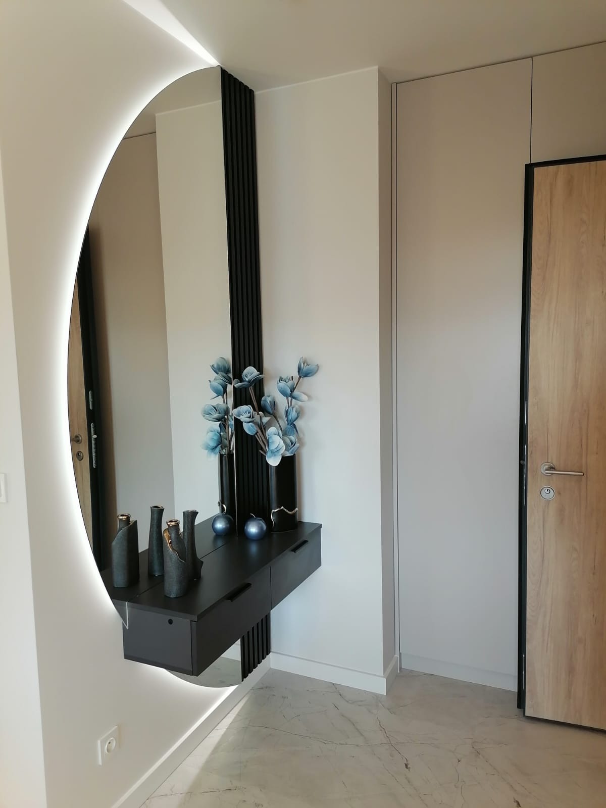 Nowoczesny przedpokój z okrągłym lustrem z podświetleniem LED, czarną konsolą z szufladami, dekoracyjnymi wazonami i kwiatami, oraz szafą wnękową z drewnianymi drzwiami.
