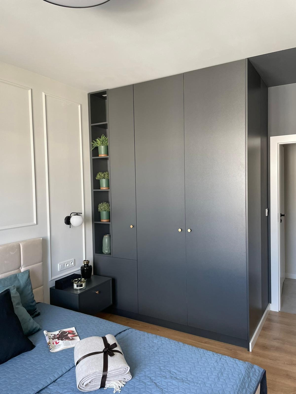 Szara szafa wnękowa z otwartymi półkami, na których stoją doniczki z roślinami, w sypialni z łóżkiem i szafką nocną, minimalistyczny design.