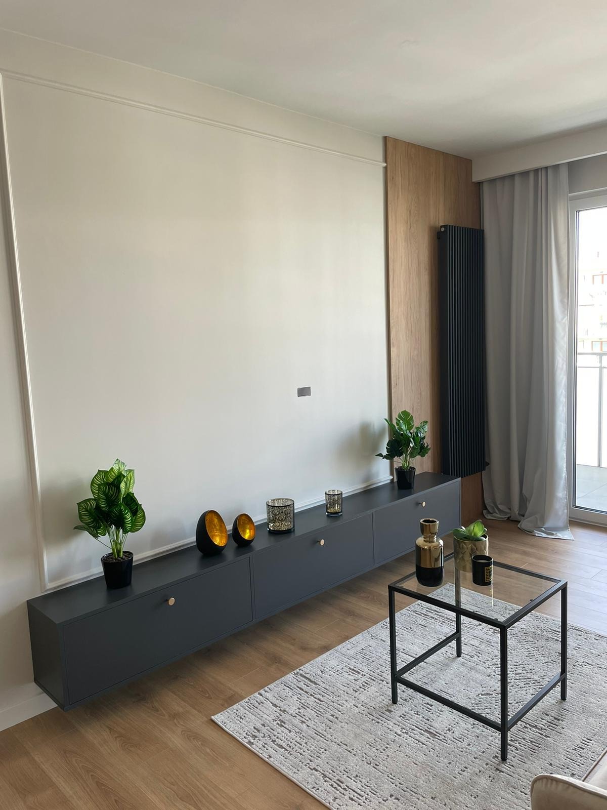 Nowoczesny salon z szarą, podwieszaną szafką RTV z dekoracjami, drewnianą ścianą i czarnym grzejnikiem.