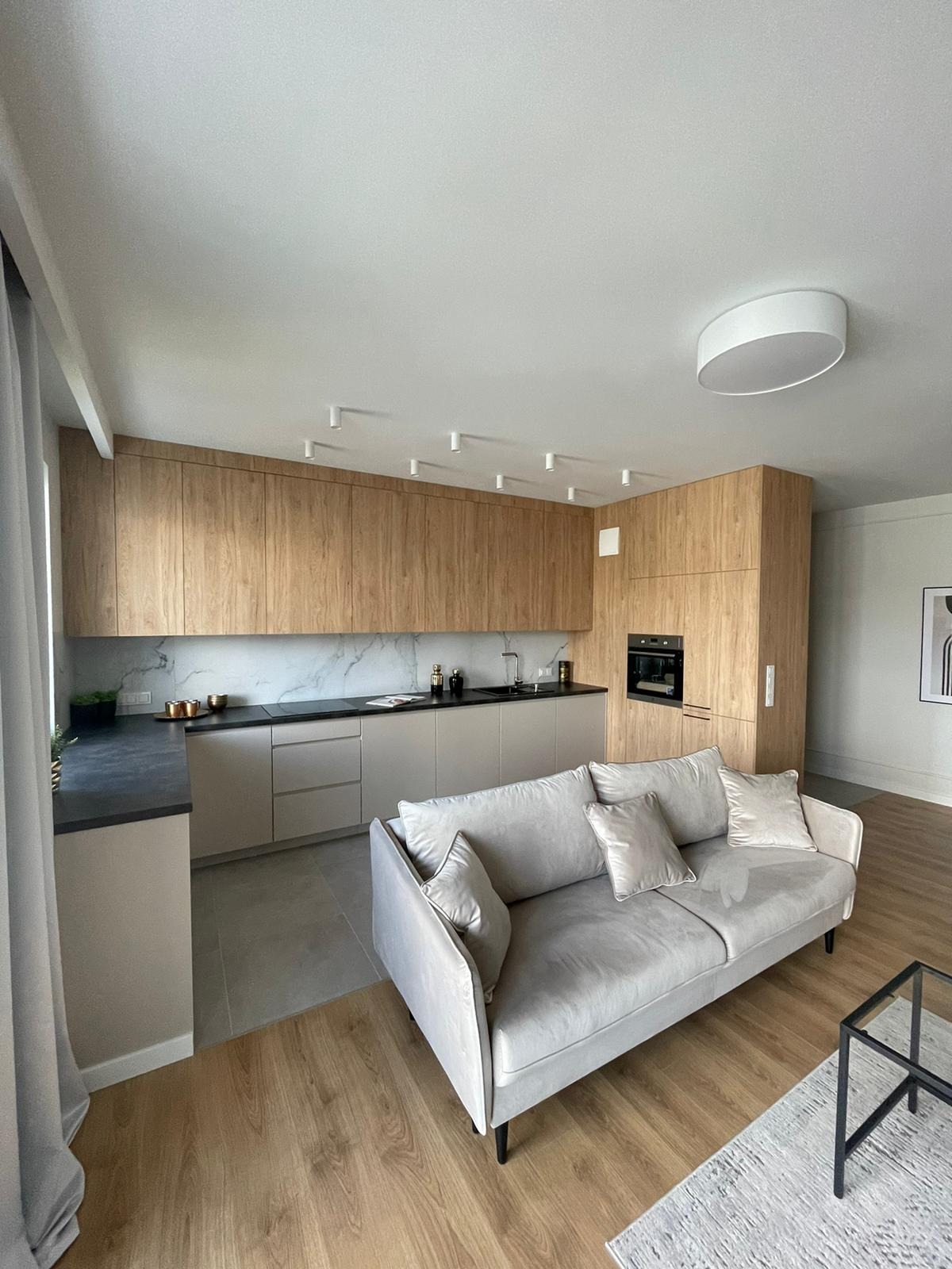 Nowoczesna kuchnia z drewnianymi frontami, szarym blatem i marmurowym backsplashiem widziana z perspektywy salonu z beżową sofą.