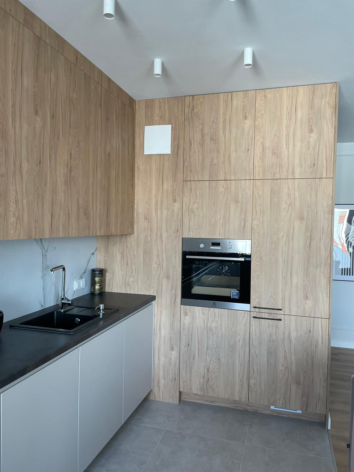 Nowoczesna kuchnia z drewnopodobnymi frontami, czarnym blatem i zlewem, szarymi szafkami dolnymi oraz wbudowanym piekarnikiem.