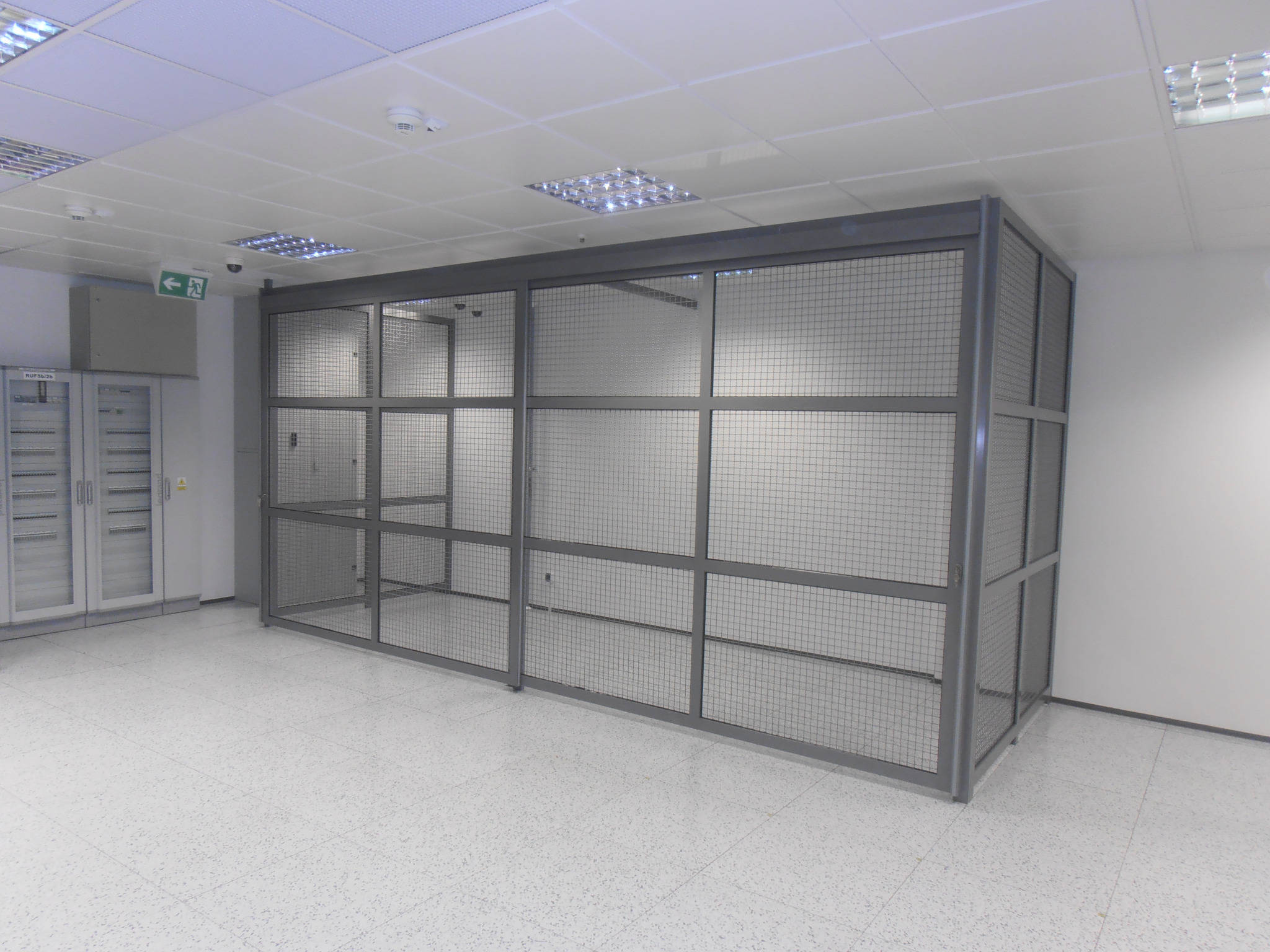 Metalowa klatka z siatki w jasnym biurze, widoczna szafa serwerowa z boku i znak ewakuacyjny.