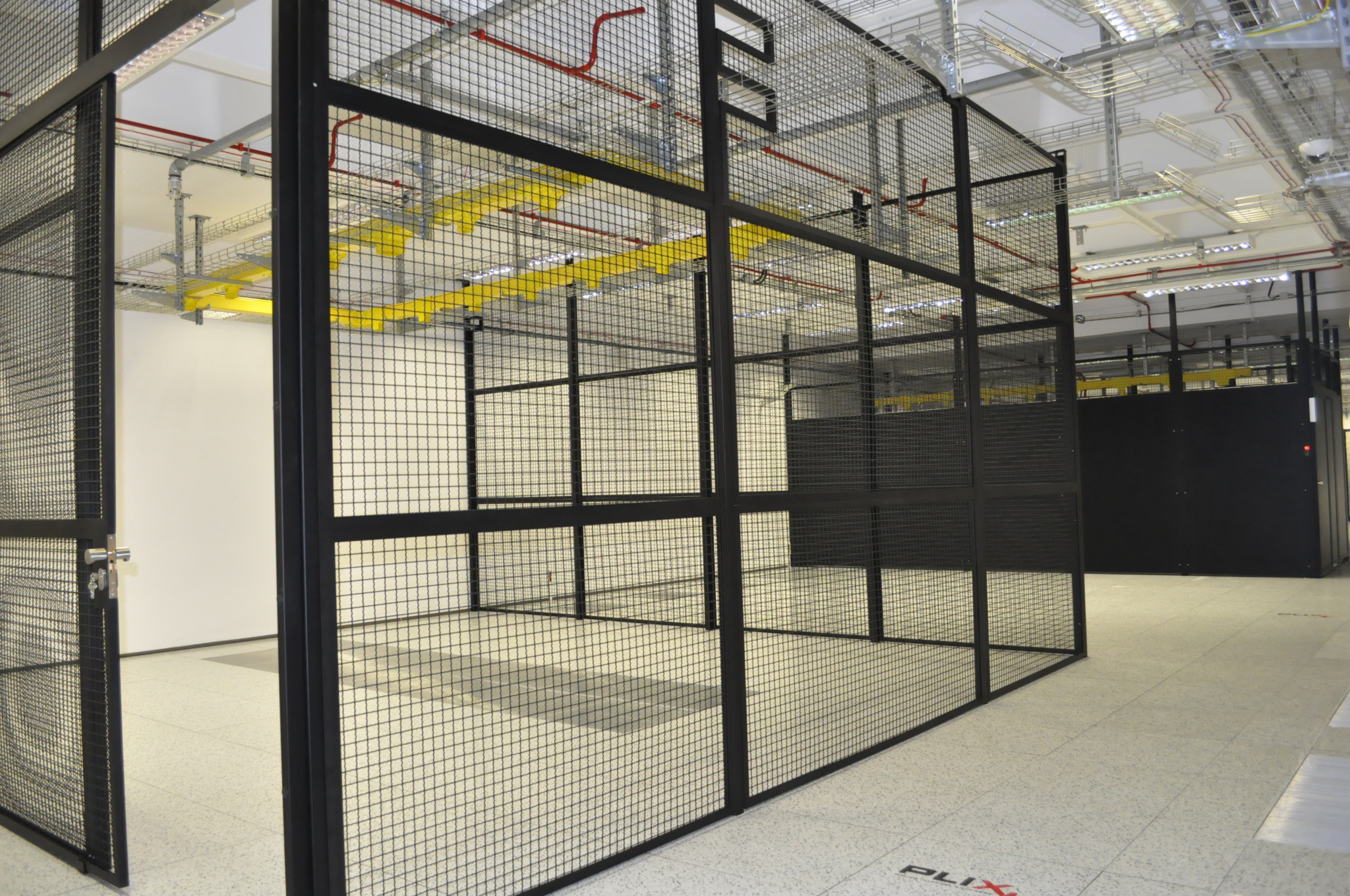 Klatka bezpieczeństwa z metalowej siatki w pomieszczeniu biurowym, widoczne instalacje pod sufitem i otwarte drzwi.